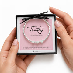 Silver bracelet with heart charms in a gift box labeled 'Thirty Happy Birthday' held by hands.