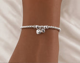 A silver beaded bracelet with two small dangling heart charms, shown worn on a person's wrist against a soft white fabric background.