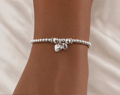 A silver beaded bracelet with two small dangling heart charms, shown worn on a person's wrist against a soft white fabric background.