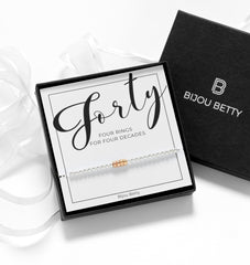 Close up of Bijou Betty’s decades bracelet gift set, 925 silver and rose gold rings, in gift box with bag, tissue, and card: “Forty - 4 rings for 4 decades.
