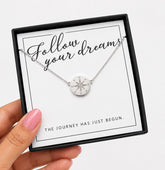 A silver compass pendant necklace on a delicate chain, presented inside a black gift box with a white card that reads 'Follow your dreams' and 'THE JOURNEY HAS JUST BEGUN.' A hand with pink nail polish holds the box against a clean white background.