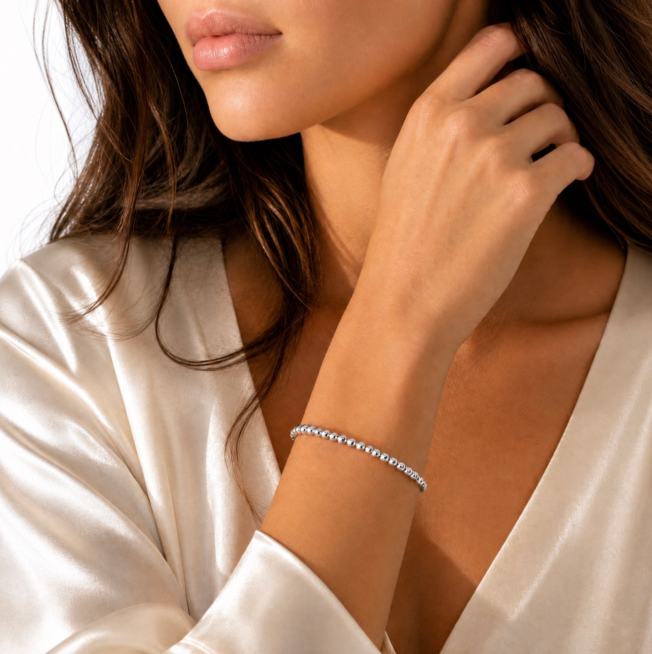 A close-up lifestyle shot of a woman wearing a delicate silver beaded bracelet on her wrist, paired with a satin cream robe.