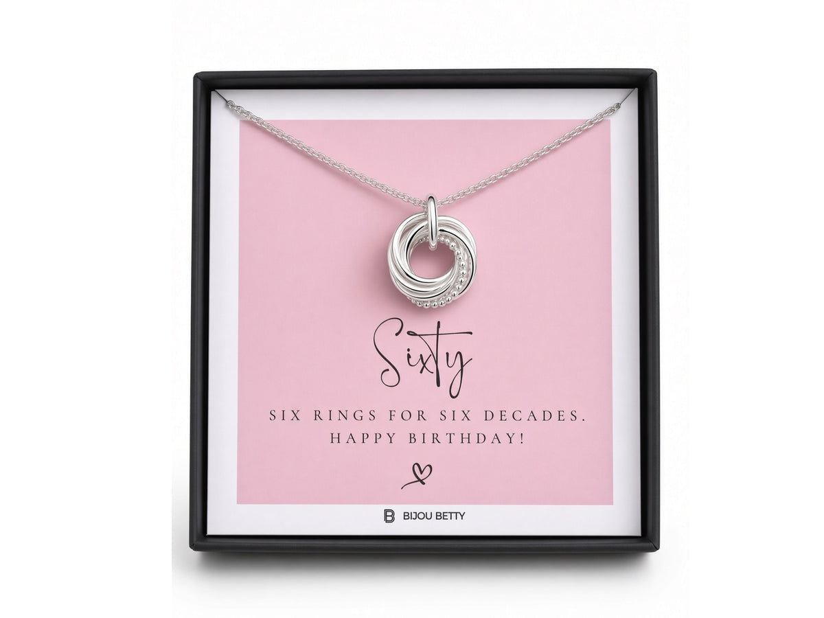 A silver ring pendant displayed inside a black gift box on a pink card that reads 'Sixty' and 'Six Rings for Six Decades. Happy Birthday!'.