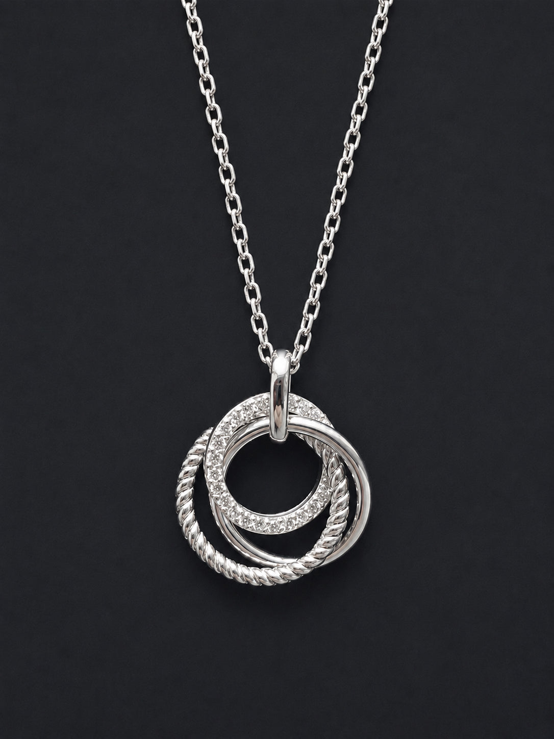 A silver 21st  pendant necklace featuring three interlocking rings, with two of the rings set with small clear stones and one twisted rope-style ring, displayed on a dark background.
