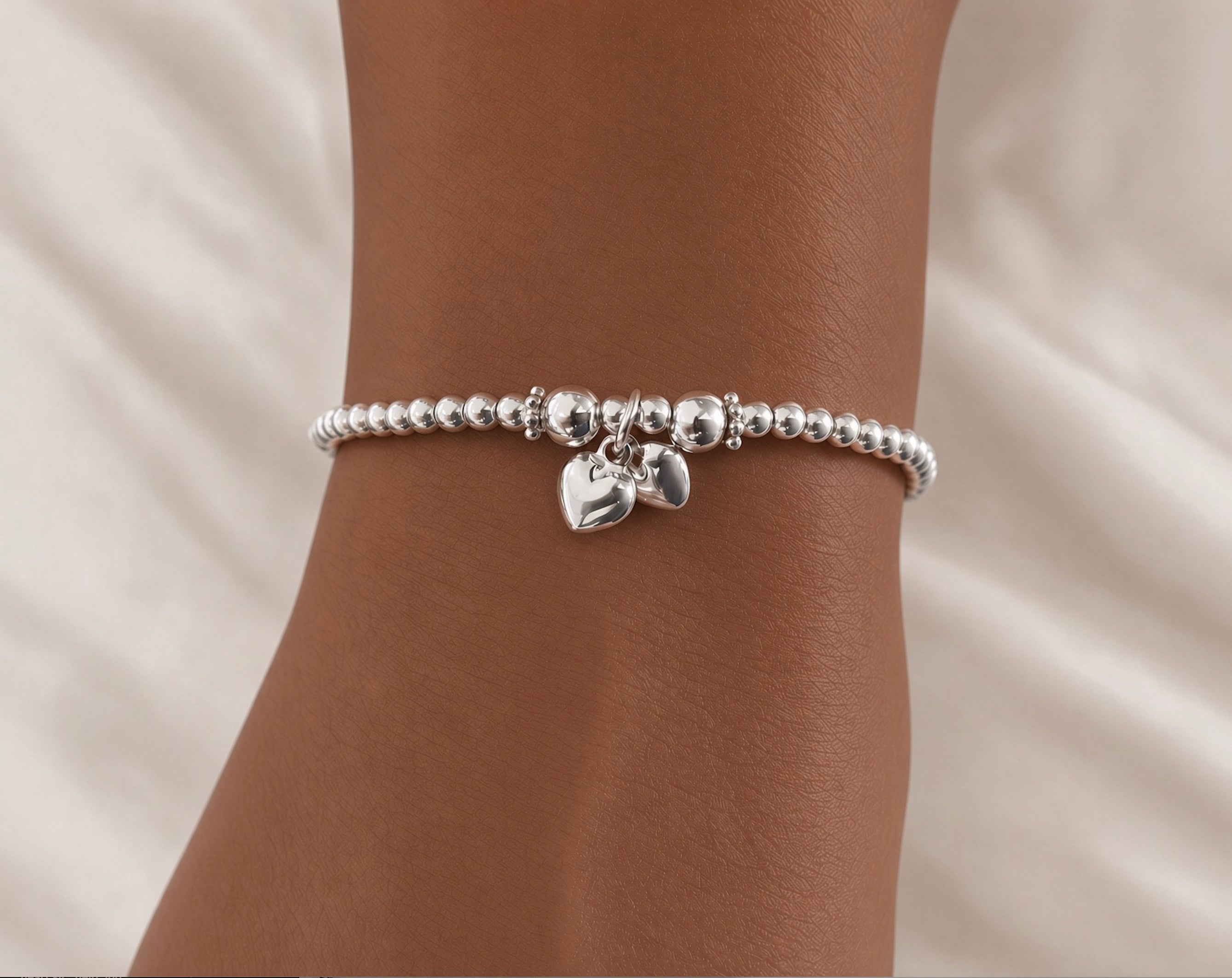A silver beaded bracelet with two small dangling heart charms, shown worn on a person's wrist against a soft white fabric background.