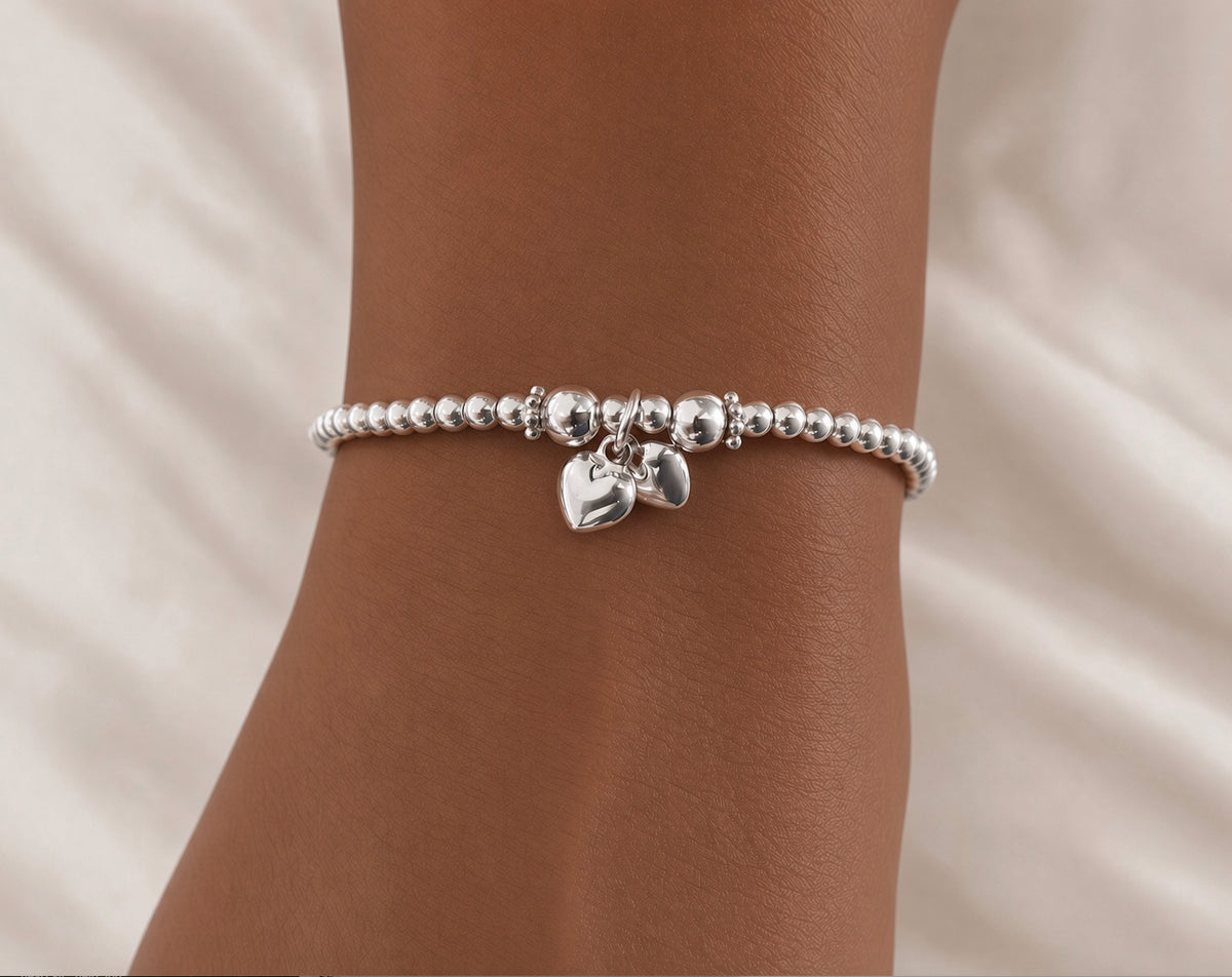 A silver beaded bracelet with two small dangling heart charms, shown worn on a person's wrist against a soft white fabric background.
