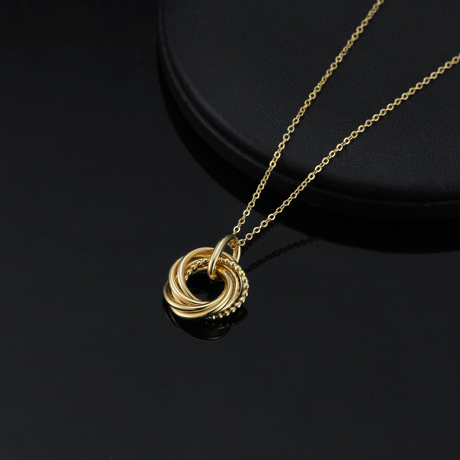 A gold necklace with a pendant featuring a circle of 5 interlocked rings. The pendant has a textured, beaded design.