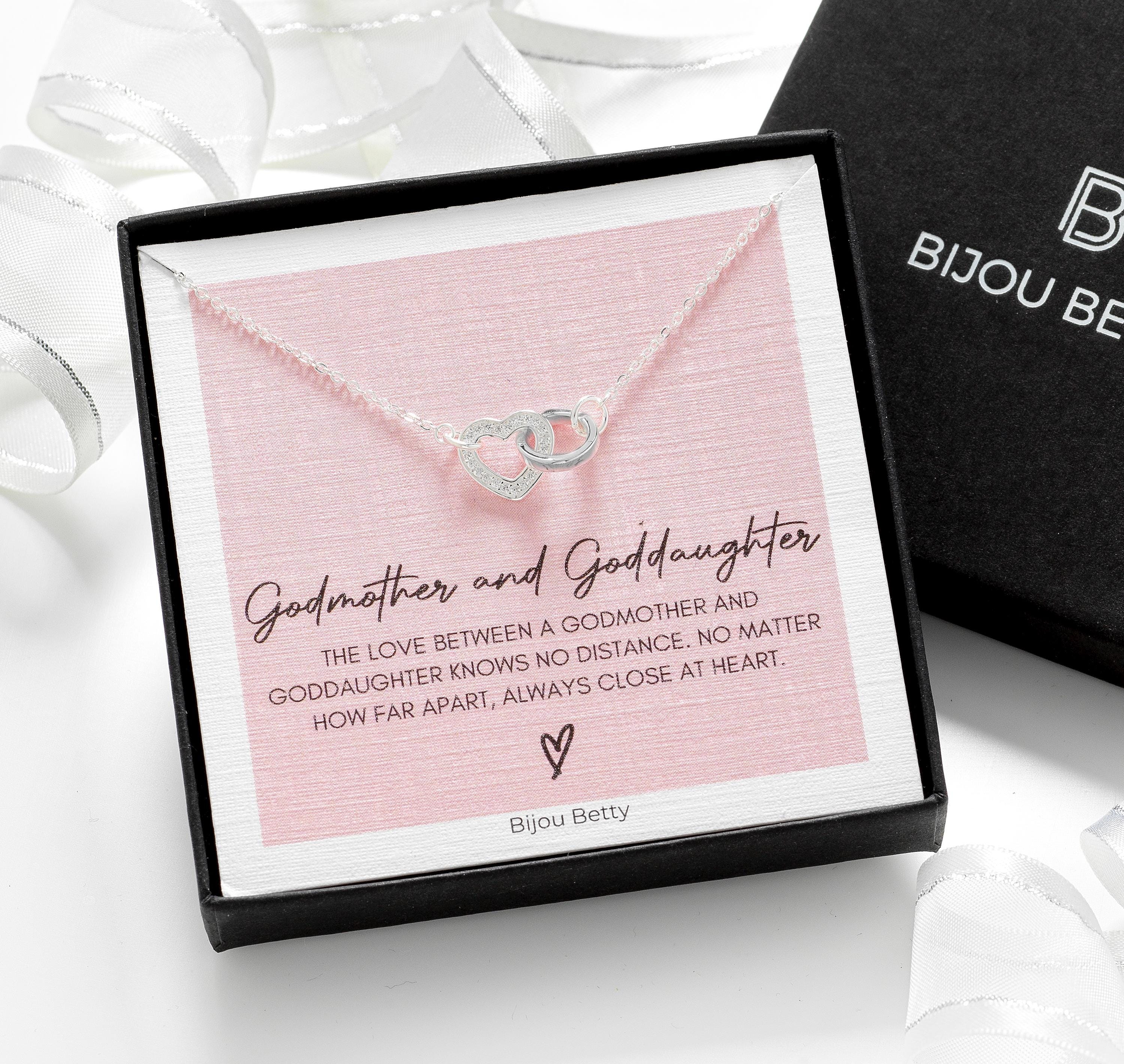 A silver necklace with two interlocking heart charms presented in a black gift box. The box features the text "Godmother & Goddaughter" and "BIJOU BETTY". The necklace is accompanied by a card that reads "The love between a godmother and goddaughter knows no distance. No matter how far apart, always close at heart.