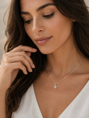 A close-up lifestyle shot of a woman wearing a delicate silver necklace with a small heart-shaped pendant, styled with a white V-neck top.