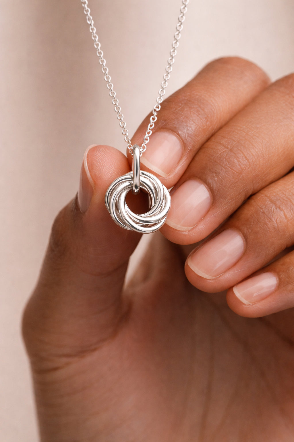 A close-up of a hand holding a silver pendant necklace with a multi-looped, knot-like design on a delicate silver chain, set against a soft beige background.