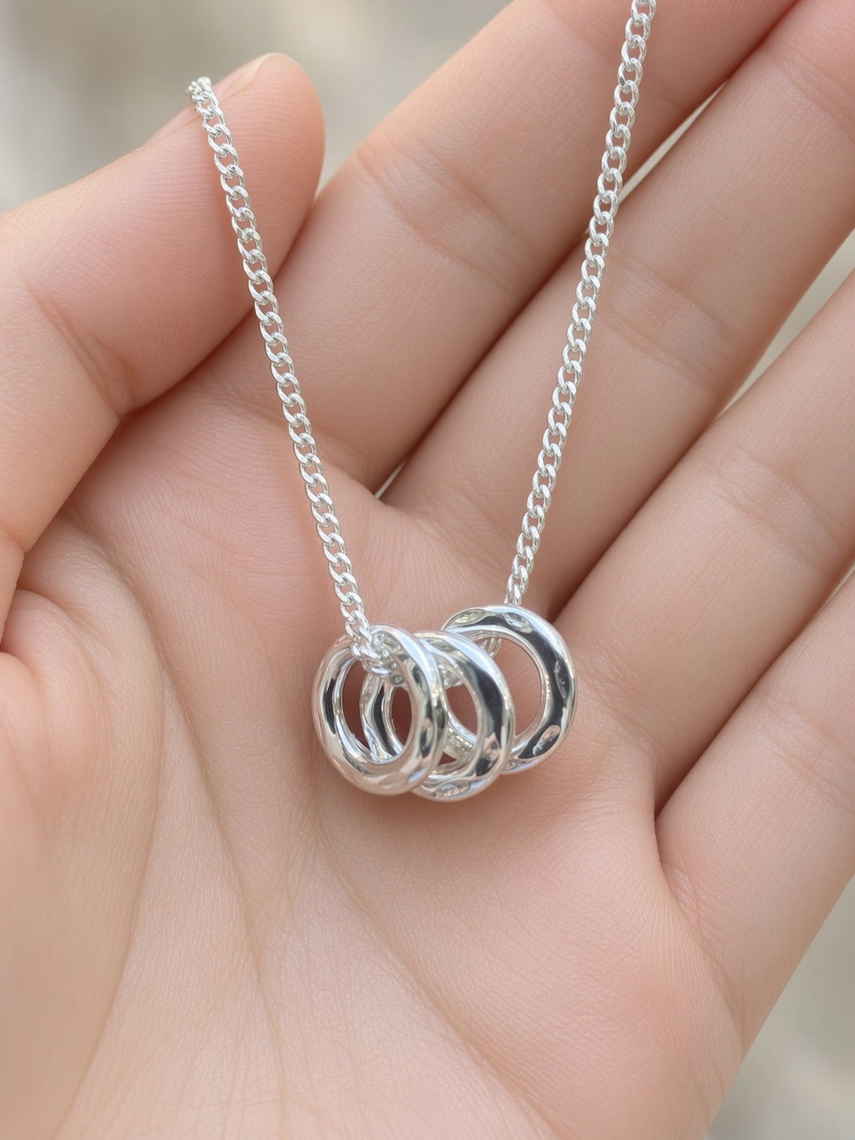 A close-up of a hand holding a silver chain necklace with three interlocking polished rings pendant.